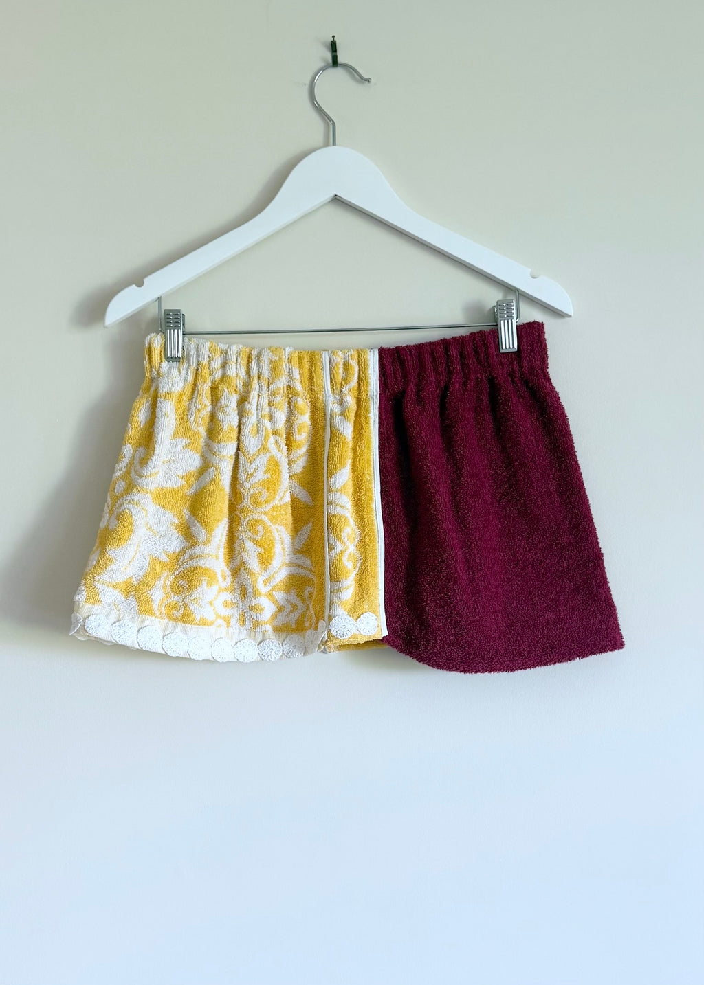 Two pairs of shorts, one yellow with white pattern and one burgundy, hanging on a white hanger against a plain background.