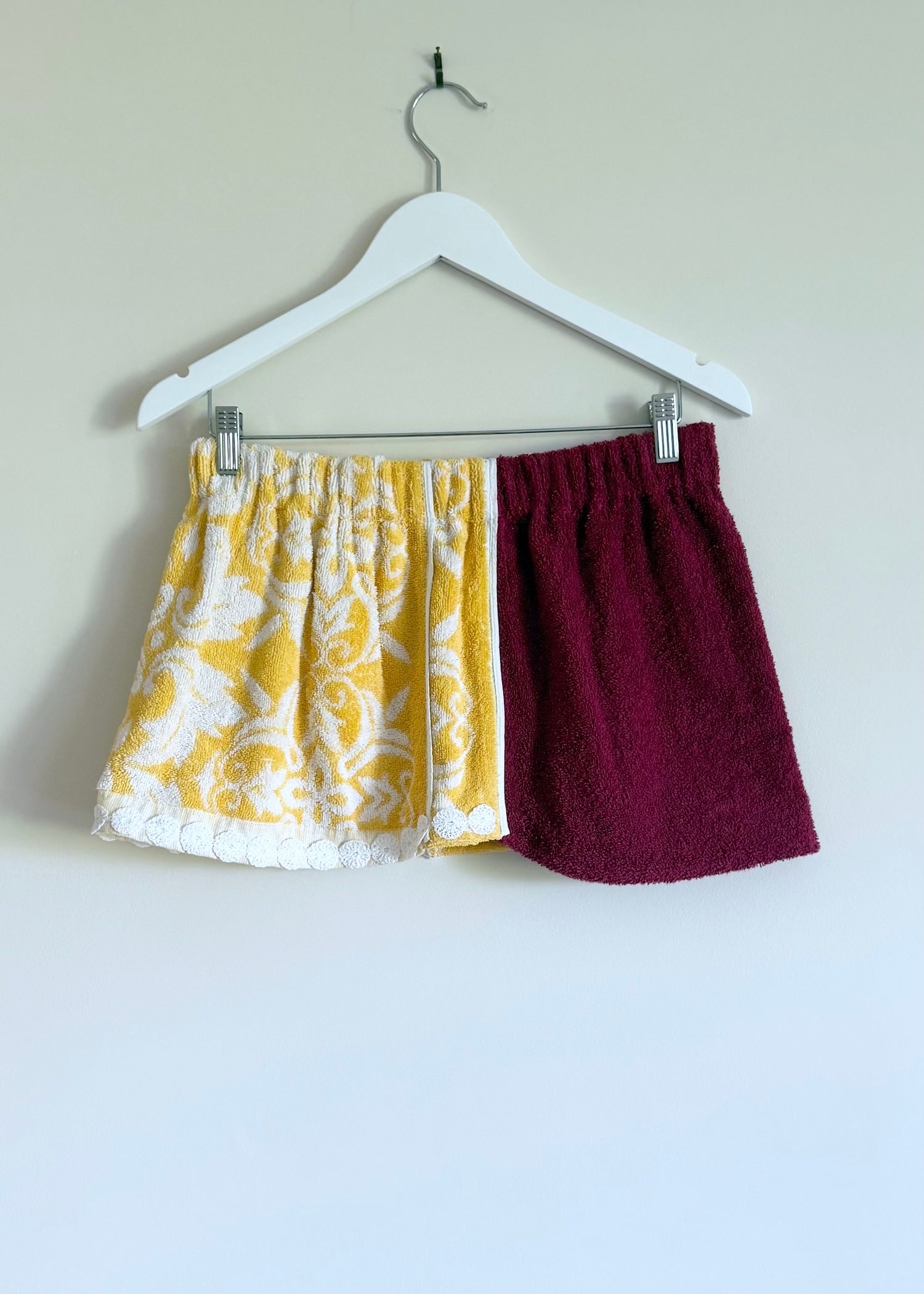 Two pairs of shorts, one yellow with white pattern and one burgundy, hanging on a white hanger against a plain background.